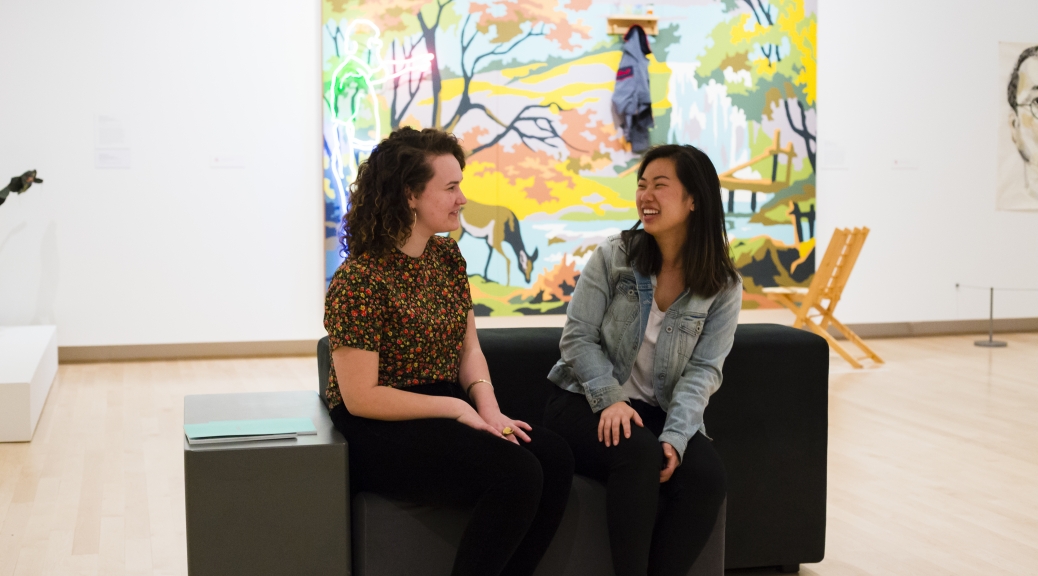 Two visitors chatting while sitting on a bench in the gallery. Behind them is a colorful work of art hanging on the wall.