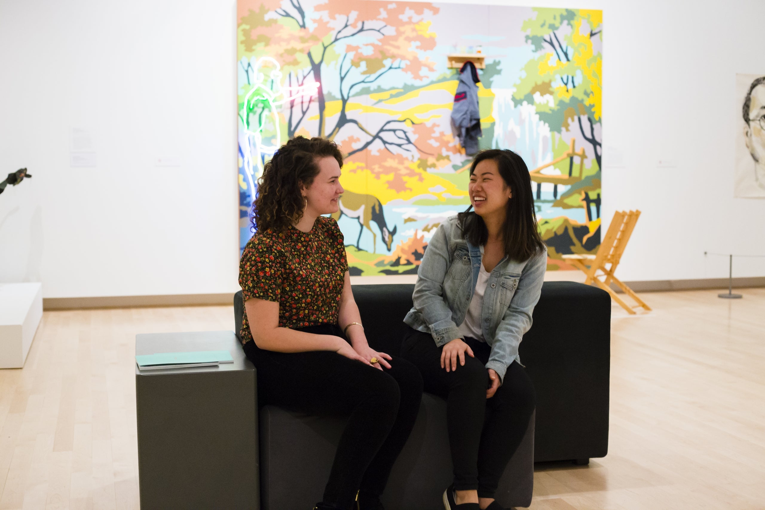 Two visitors chatting while sitting on a bench in the gallery. Behind them is a colorful work of art hanging on the wall.