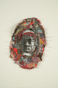 Small brooch-like sculpture with a photo of a young African American girl at center surrounded by a border of wire and cloth fragments.