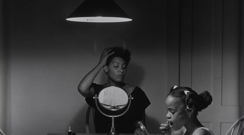 A black and white photography taken by artist Carrie Mae Weems that features a black woman sits at the kitchen table styling her hair while her daughter puts on lipstick next to her.