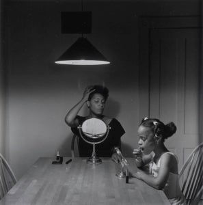 A black and white photography taken by artist Carrie Mae Weems that features a black woman sits at the kitchen table styling her hair while her daughter puts on lipstick next to her.