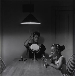 A black and white photography taken by artist Carrie Mae Weems that features a black woman sits at the kitchen table styling her hair while her daughter puts on lipstick next to her.