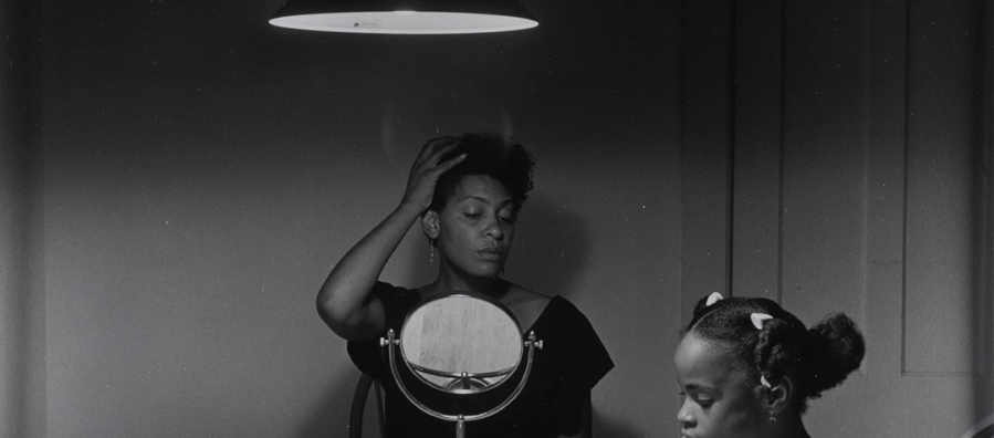 A black and white photography taken by artist Carrie Mae Weems that features a black woman sits at the kitchen table styling her hair while her daughter puts on lipstick next to her.