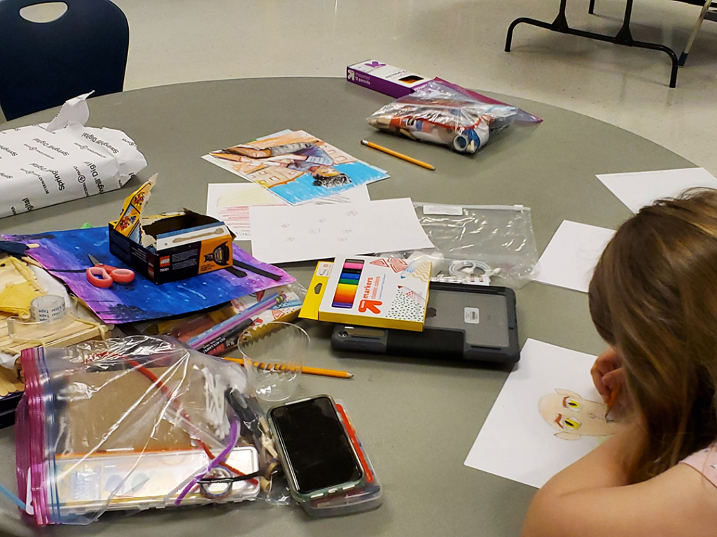 Child drawing amongst art supplies strewn over a table. 