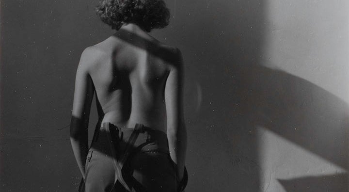 A woman in trousers and suspenders stands facing a wall with her back to the viewer. She is nude from the waist up.