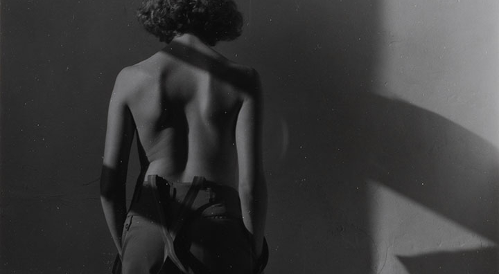 A woman in trousers and suspenders stands facing a wall with her back to the viewer. She is nude from the waist up.