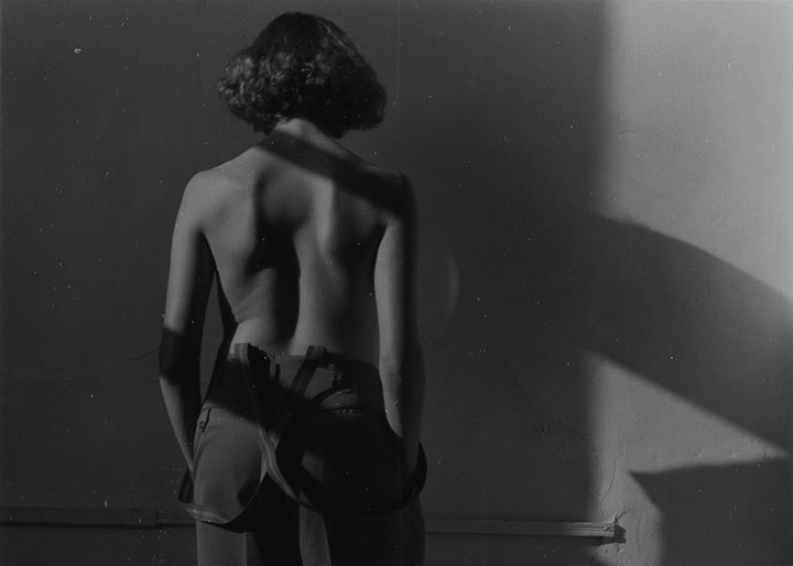 A woman in trousers and suspenders stands facing a wall with her back to the viewer. She is nude from the waist up.