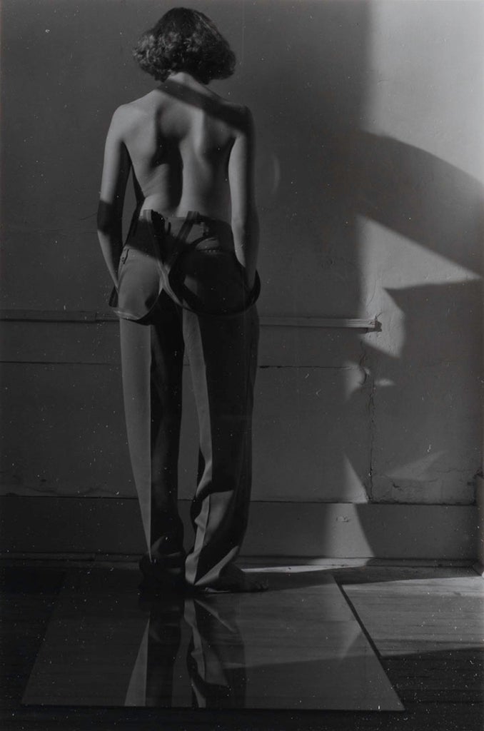 A woman in trousers and suspenders stands facing a wall with her back to the viewer. She is nude from the waist up.