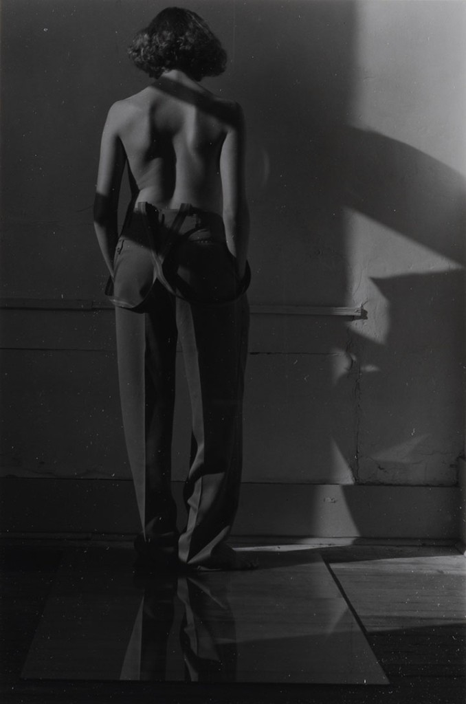 A woman in trousers and suspenders stands facing a wall with her back to the viewer. She is nude from the waist up.