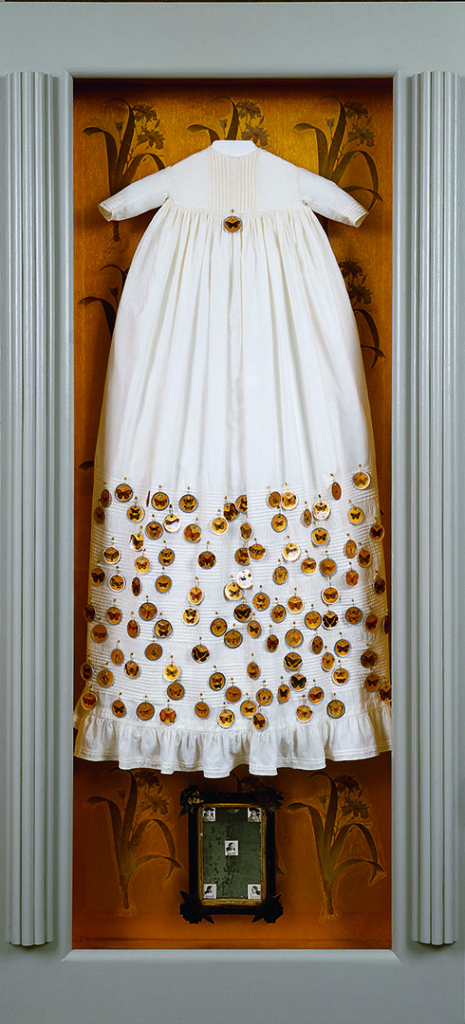 Gold painted background with flower pattern. Cream antique infant dress with round gold butterfly tokens safety pinned to skirt of dress. One token on breast of dress. Below dress is a picture frame 7" x 9" with five small gelatin silver prints of a young girl in the corners and center of the frame.