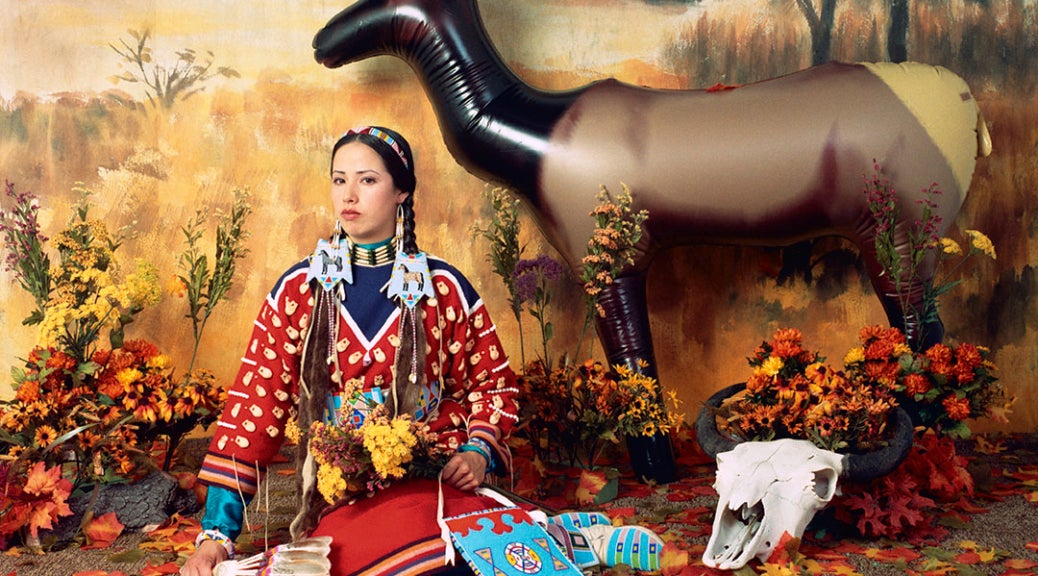 Staged diorama-like image of a woman in a colorful skirt and blouse, wearing elaborate jewelry and hair ornaments holding a feather fan, wildflowers and a beaded bag seated in front of a backdrop painted with an autumn landscape. Leaves scattered on the ground, a plastic elk and an animal skull to her left.