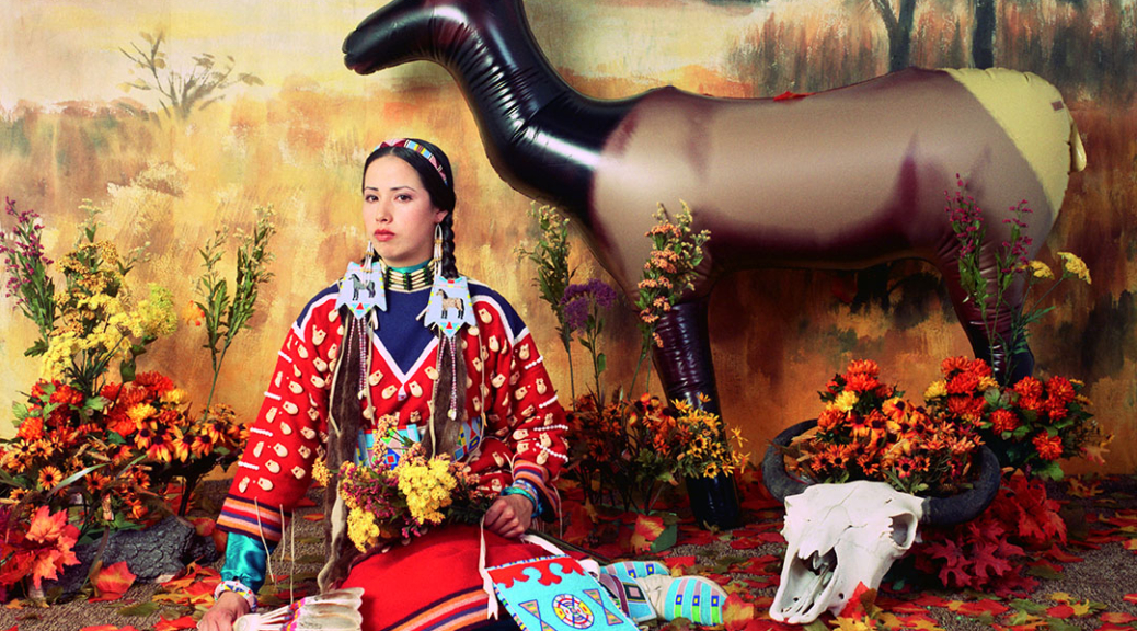 Staged diorama-like image of a woman in a colorful skirt and blouse, wearing elaborate jewelry and hair ornaments holding a feather fan, wildflowers and a beaded bag seated in front of a backdrop painted with an autumn landscape. Leaves scattered on the ground, a plastic elk and an animal skull to her left.