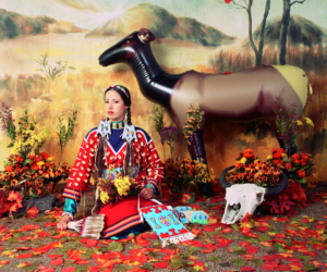 Staged diorama-like image of a woman in a colorful skirt and blouse, wearing elaborate jewelry and hair ornaments holding a feather fan, wildflowers and a beaded bag seated in front of a backdrop painted with an autumn landscape. Leaves scattered on the ground, a plastic elk and an animal skull to her left.