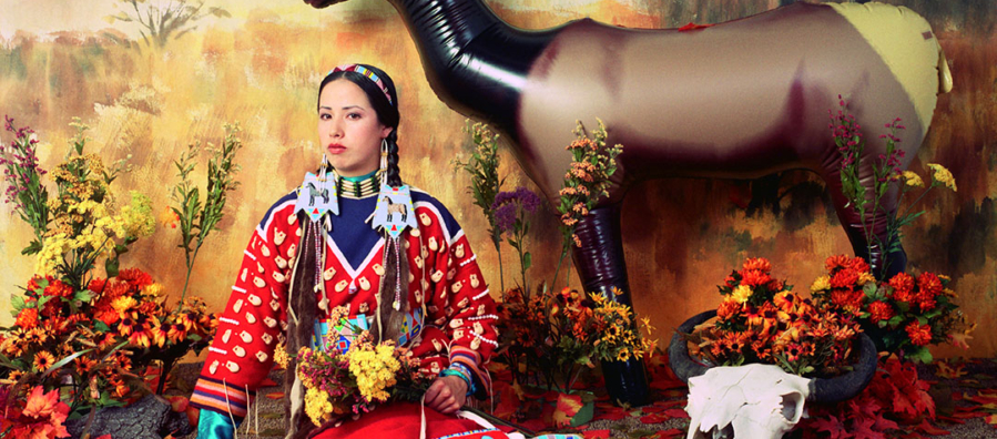 Staged diorama-like image of a woman in a colorful skirt and blouse, wearing elaborate jewelry and hair ornaments holding a feather fan, wildflowers and a beaded bag seated in front of a backdrop painted with an autumn landscape. Leaves scattered on the ground, a plastic elk and an animal skull to her left.