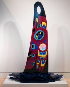 A large fin-shaped piece of glass with a red background, black top, and base made of blue metal wave shapes. The body of the "fin" is decorated with Native American designs in blue, yellow, red, black and white. A glass face is inset into the fin at lower left.