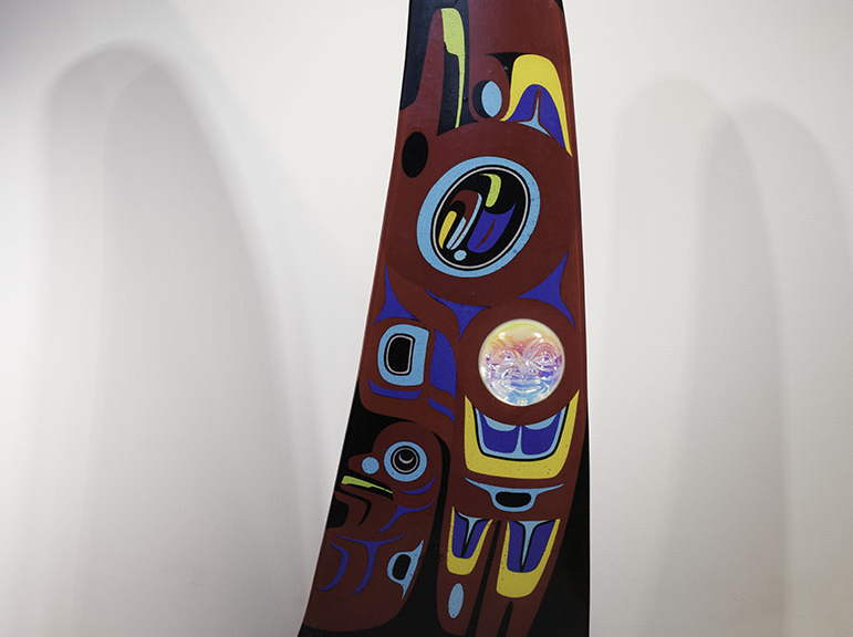 A large fin-shaped piece of glass with a red background, black top, and base made of blue metal wave shapes. The body of the "fin" is decorated with Native American designs in blue, yellow, red, black and white. A glass face is inset into the fin at lower left.