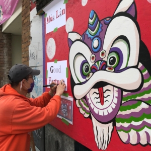 Artist Nori Kimura works on his mural depicting a colorful dragon on a vibrant red background.