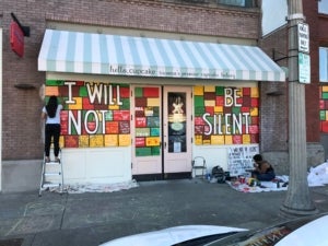 Two figures at work painting a mural composed of mulitcolored squares with small text. Over the squares, text in white reads "I will not be silent."