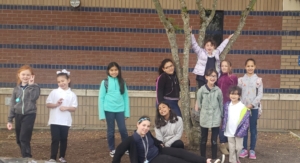 Eleven grade school students stand outside of the school around a tree. 