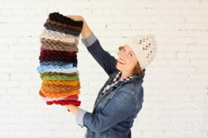 Woman wearing a denim jacket and a knit white hat holds a multicolored stack of knit products.