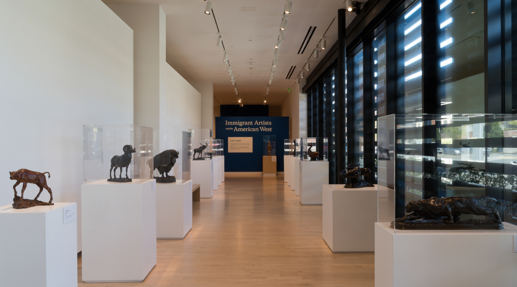 Long corridor shaped gallery with wooden floors and white walls. Pedestals punctuate the sides of the gallery holding bronze sculptures. A blue wall at the end of the gallery reads Immigrant Artists and the American West.