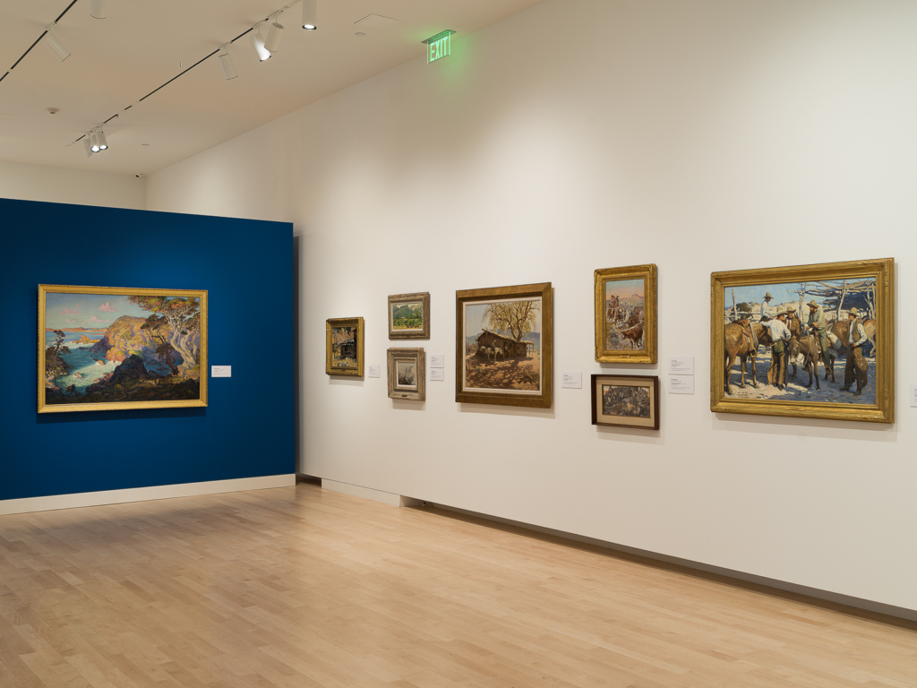 View from Immigrant Artists and the American West featuring 7 paintings hung on a white wall. A bright blue wall with a single painting is perpendicular to the white wall.