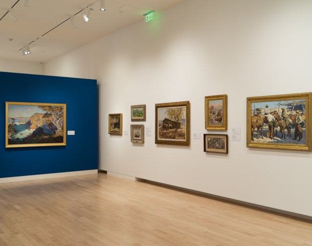 View from Immigrant Artists and the American West featuring 7 paintings hung on a white wall. A bright blue wall with a single painting is perpendicular to the white wall.