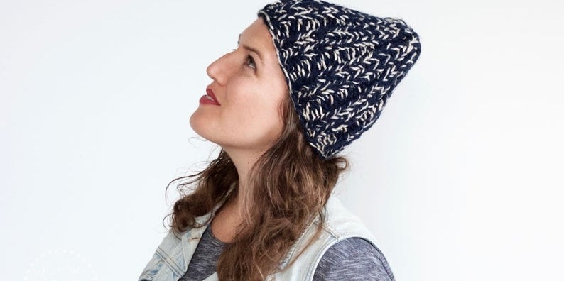 Anna Sharp, owner of Victory Garden Yard, looks up toward the sky wearing a hand knit blue and white beanie.