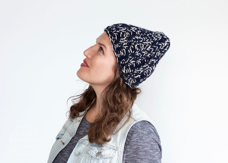 Anna Sharp, owner of Victory Garden Yard, looks up toward the sky wearing a hand knit blue and white beanie.