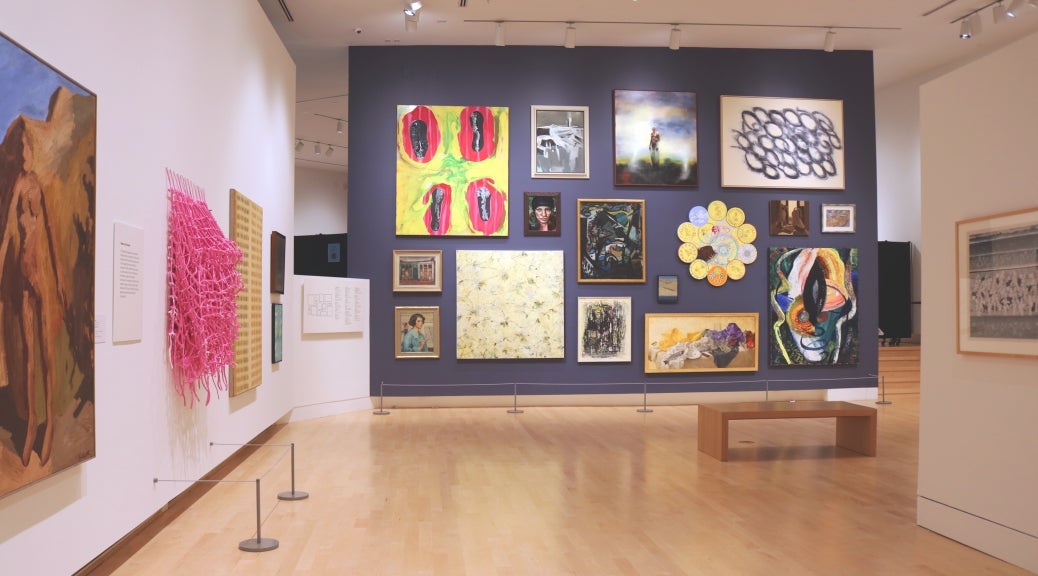Photograph of a museum gallery space with a salon-style wall where 16 paintings are hung on a dark grey wall.