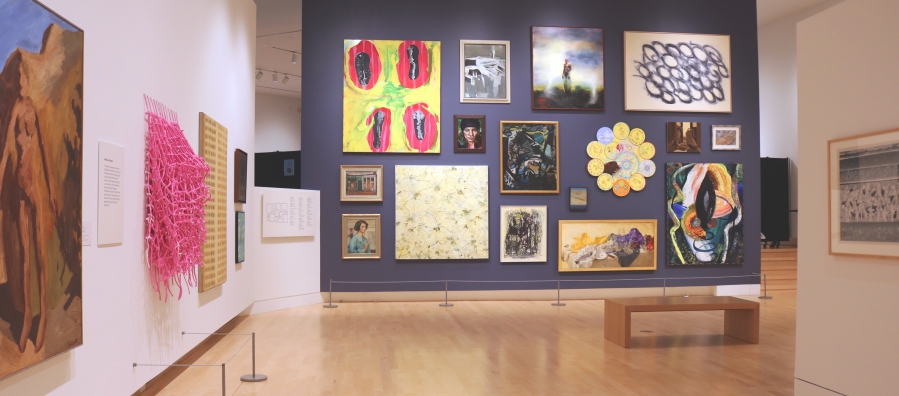 Photograph of a museum gallery space with a salon-style wall where 16 paintings are hung on a dark grey wall.