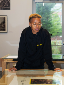 Color photo of a person wearing a black and yellow hooded sweater leaned over a table.