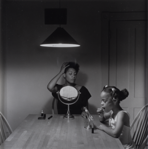 Black and white photograph of two figures applying makeup at a dining room table.