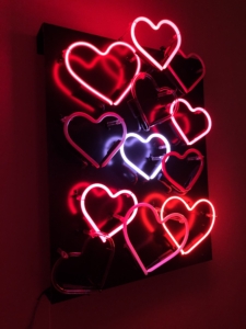 Color photo of neon hearts in various shades of pink and red set against a black background.