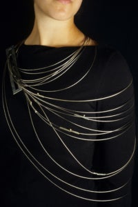 Photograph of a person in a black sweater wearing a silver body piece made up of cords of various lengths wrapped around the body.