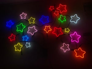 Color photo of neon stars lit up in various colors and hanging on a black wall.