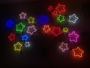 Color photo of neon stars lit up in various colors and hanging on a black wall.